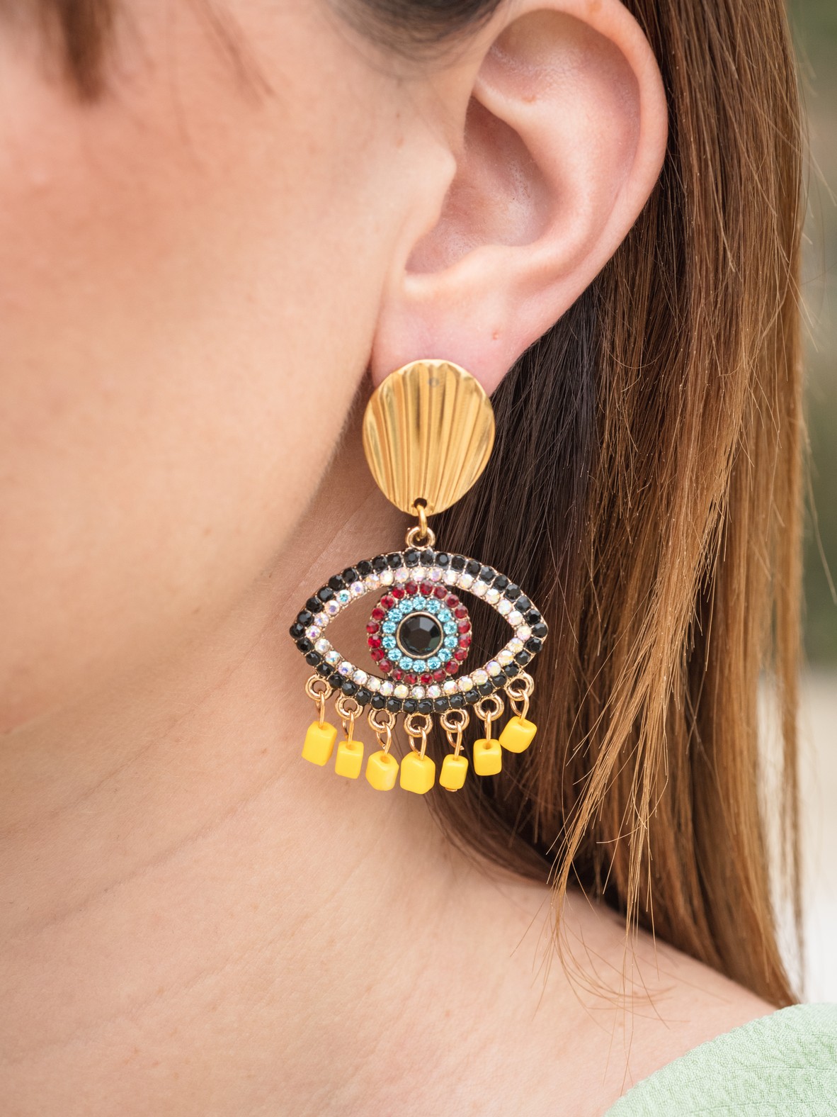 Statement σκουλαρίκια με μεταλλικό στοιχείο-μάτι διακοσμημένο με πολύχρωμα στρας και έντονη χρωματική αντίθεση σε μαύρο, κόκκινο, γαλάζιο και κίτρινο. Το πάνω μέρος θυμίζει χρυσό κοχύλι, ενώ η βάση του ματιού κοσμείται με κίτρινες κρεμαστές λεπτομέρειες για παιχνιδιάρικη κίνηση. Διαθέτουν σταθερό ατσάλινο κούμπωμα για άνετη και ασφαλή εφαρμογή.