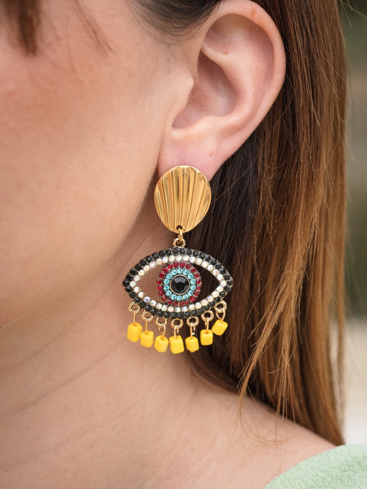 Statement σκουλαρίκια με μεταλλικό στοιχείο-μάτι διακοσμημένο με πολύχρωμα στρας και έντονη χρωματική αντίθεση σε μαύρο, κόκκινο, γαλάζιο και κίτρινο. Το πάνω μέρος θυμίζει χρυσό κοχύλι, ενώ η βάση του ματιού κοσμείται με κίτρινες κρεμαστές λεπτομέρειες για παιχνιδιάρικη κίνηση. Διαθέτουν σταθερό ατσάλινο κούμπωμα για άνετη και ασφαλή εφαρμογή.