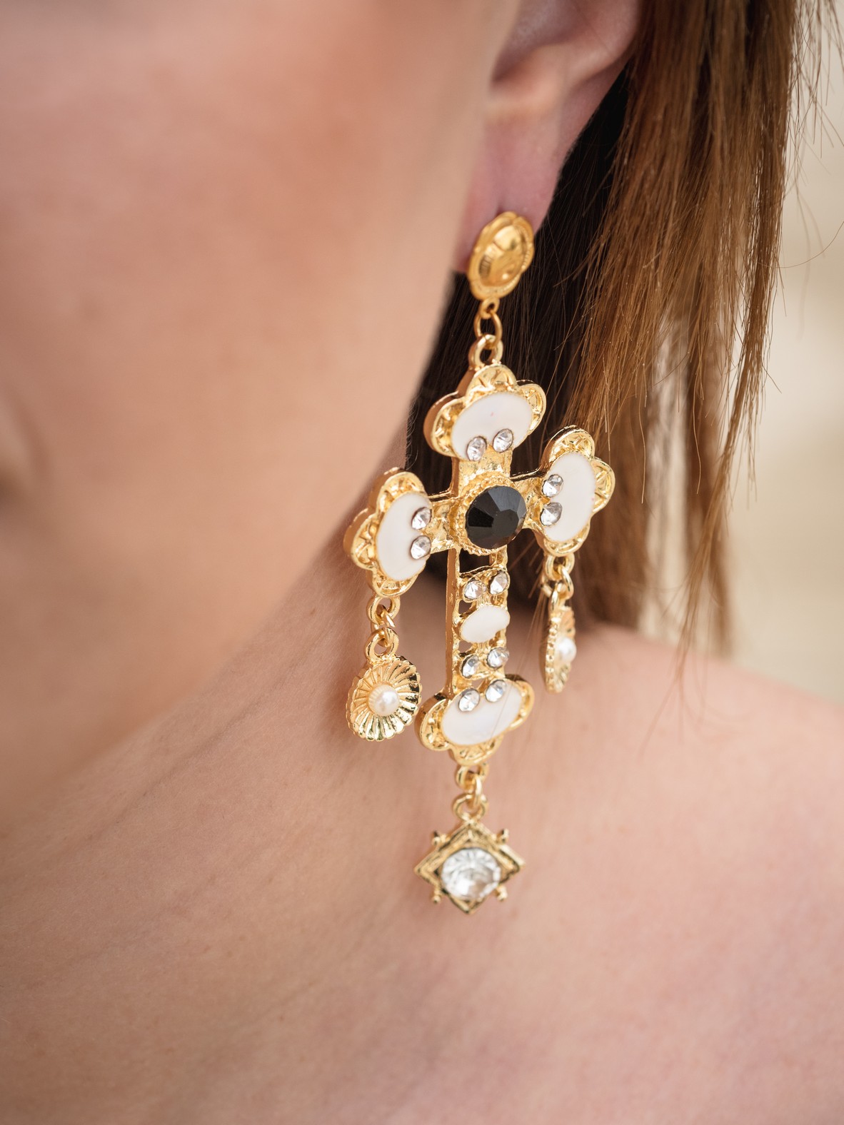 Εντυπωσιακά σκουλαρίκια σε σχέδιο σταυρού, με ανάγλυφες λεπτομέρειες και έντονη χρωματική αντίθεση σε χρυσό, λευκό και μαύρο. Το μεταλλικό στοιχείο στο κέντρο αναδεικνύεται από την μαύρη πέτρα, ενώ τα διακοσμητικά στρας και οι κρεμαστές λεπτομέρειες προσθέτουν πολυτέλεια και λάμψη. Στηρίζονται σε ατσάλινο κούμπωμα για άνεση και σταθερότητα.