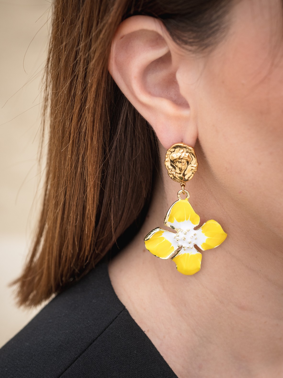 Ζωντανά και κομψά σκουλαρίκια με επιχρυσωμένη βάση και εντυπωσιακό λουλούδι σε έντονο κίτρινο και λευκό σμάλτο. Το επάνω μέρος διακοσμείται με ανάγλυφο πρόσωπο, προσθέτοντας μια καλλιτεχνική πινελιά και μοναδικότητα στο σχέδιο. Ελαφριά και ευκολοφόρετα, ιδανικά για να δώσουν χρώμα και χαρακτήρα στις ανοιξιάτικες και καλοκαιρινές σου εμφανίσεις.