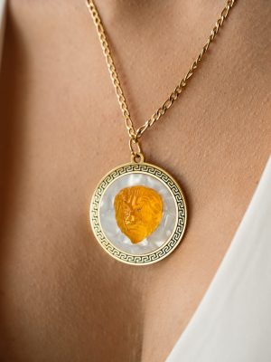 Χρυσό μενταγίον με ανάγλυφο κεφάλι λιονταριού σε κεχριμπαρένια απόχρωση, τοποθετημένο πάνω σε φόντο περλέ, και πλαισιωμένο με κλασικό μοτίβο μαίανδρο.