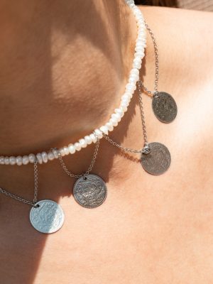 Κομψό choker που συνδυάζει την καθαρότητα των λευκών περλών με τη γοητεία των ατσάλινων στοιχείων σε μορφή αρχαΐκων νομισμάτων. Το σχέδιο αντλεί έμπνευση από τα παλιά ελληνικά φυλαχτά και τα κοσμήματα εποχής.