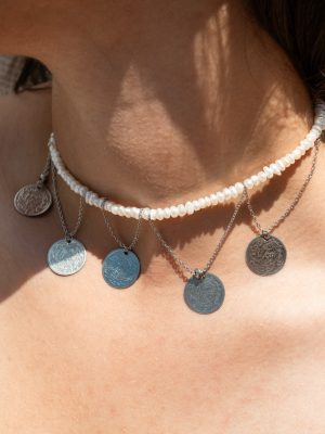 Κομψό choker που συνδυάζει την καθαρότητα των λευκών περλών με τη γοητεία των ατσάλινων στοιχείων σε μορφή αρχαΐκων νομισμάτων. Το σχέδιο αντλεί έμπνευση από τα παλιά ελληνικά φυλαχτά και τα κοσμήματα εποχής.